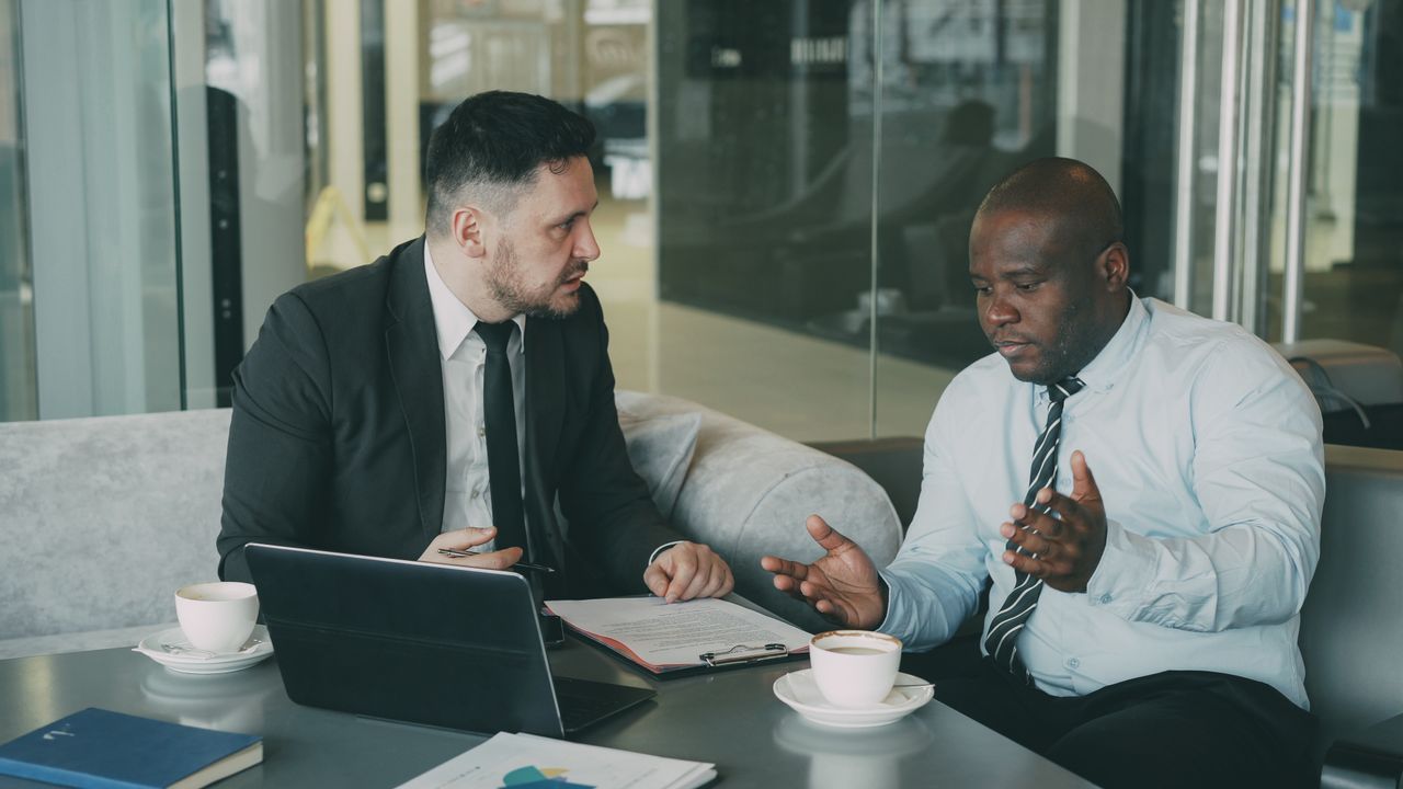 Mastering the First Impression: Your intriguing post title goes here Two businessmen in discussion over coffee and laptop in modern office setting.